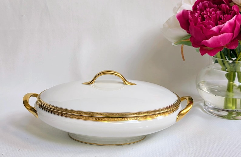 May include: A white porcelain serving dish with a lid, featuring a gold-toned handle and two gold-toned side handles. The rim of the lid and the base of the dish are decorated with a detailed gold band pattern. The dish has a smooth finish.