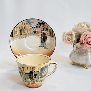 May include: A vintage teacup and saucer set with a street scene design. The cup and saucer feature a yellow and orange gradient, with a black rim. A small vase with pink and white roses is to the right.