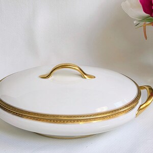 May include: A white porcelain serving dish with a lid, featuring a gold-toned handle on top and two gold-toned side handles. The rim of the dish is decorated with a detailed gold band featuring a repeating geometric pattern around the edge.