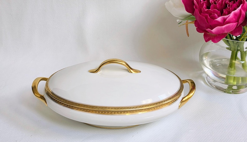 May include: A white porcelain serving dish with a lid, featuring a gold-toned handle on top and two gold-toned side handles. The rim of the dish is decorated with a detailed gold band featuring a repeating geometric pattern around the edge.