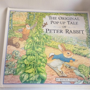 以下が含まれることがあります： ビアトリクス・ポター著『The Original Pop-Up Tale of Peter Rabbit』というタイトルのハードカバー本。表紙には、青いジャケットを着たウサギが庭を駆け抜け、背景で男性が手押し車を押しているカラフルなイラストが描かれています。