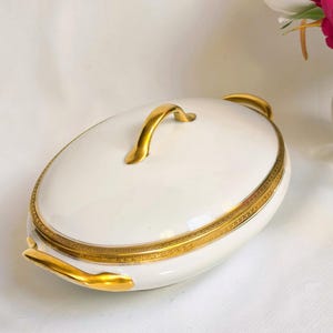 May include: An oval white porcelain serving dish with a lid, featuring a gold-toned handle on top and two gold-toned side handles. The rim of the lid and the base of the dish are decorated with a detailed, intricate gold-colored border pattern.