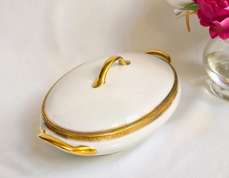 May include: An oval white porcelain serving dish with a lid, featuring a gold-toned handle on top and two gold-toned side handles. The rim of the lid and the base of the dish are decorated with a detailed, intricate gold-colored border pattern.