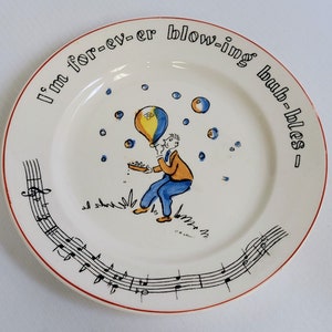 Op de afbeelding: Een wit keramiek bord met een rode rand heeft een cartoonillustratie van een persoon die bellen blaast. Het bord heeft een muzieknootontwerp rond de rand en de tekst "I'm for-ev-er blowing bub-bles".