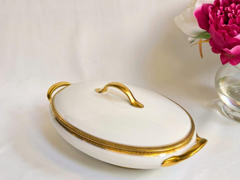 May include: An oval white porcelain serving dish with a lid, featuring a gold-toned handle and a decorative gold trim along the rim. The dish includes two gold-toned side handles, providing a classic look for serving food at the table.