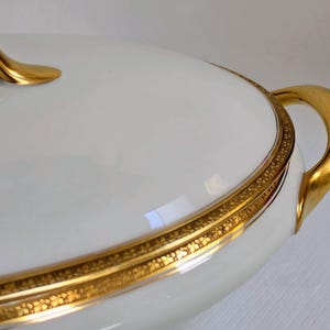 May include: A white porcelain tureen with a lid, featuring a smooth, glossy finish. The piece is accented with a wide, ornate gold band around the rim and lid edge, complemented by curved, polished gold handles on the sides and top.