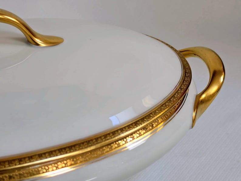 May include: A white porcelain tureen with a lid, featuring a smooth, glossy finish. The piece is accented with a wide, ornate gold band around the rim and lid edge, complemented by curved, polished gold handles on the sides and top.