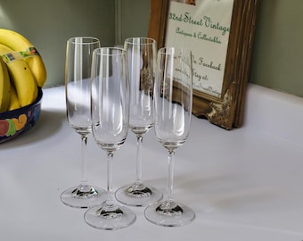 May include: Four clear champagne flutes with long stems and round bases are arranged on a white surface. The flutes are empty and reflect light. A bowl of bananas and a framed sign are in the background.