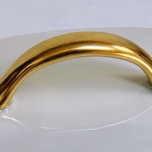 May include: A polished gold-toned metal handle with a smooth, arched design. The handle features a curved, elongated shape that tapers at both ends, securely attached to a smooth, white, curved surface that reflects the metallic gold finish.