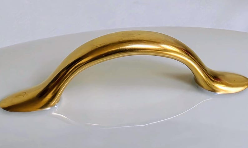 May include: A polished gold-toned metal handle with a smooth, arched design. The handle features a curved, elongated shape that tapers at both ends, securely attached to a smooth, white, curved surface that reflects the metallic gold finish.