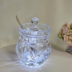 May include: A clear glass sugar bowl featuring a faceted geometric lid and a starburst cut pattern on the rounded body. A small silver-toned metal spoon rests inside the bowl, protruding through a notch in the lid. The glass reflects light.