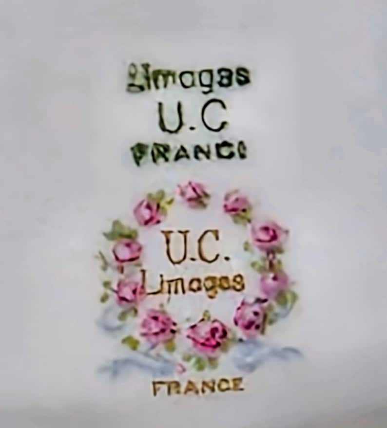 May include: A white ceramic surface featuring a maker's mark with the text Limoges U.C. FRANCE. Below this, a circular wreath of pink roses and green leaves surrounds the text U.C. Limoges, with the word FRANCE printed at the bottom.