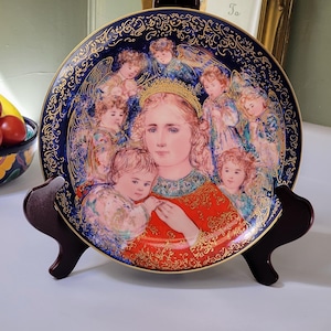 Puede incluir: Plato decorativo con una imagen central de una mujer y un niño rodeados de figuras de querubines. El plato tiene un borde azul oscuro con filigrana dorada y se exhibe en un soporte de madera. La mujer lleva una prenda roja y dorada.