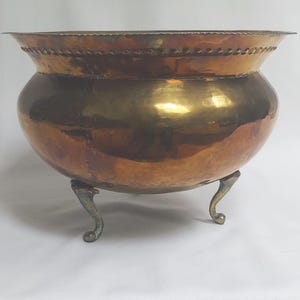 Pode incluir: Uma taça de cobre polido com corpo arredondado e uma borda larga e alargada com um detalhe decorativo de contas. A taça é suportada por três pernas de metal curvas que apresentam uma pátina ligeiramente envelhecida e escura em relação ao corpo brilhante.