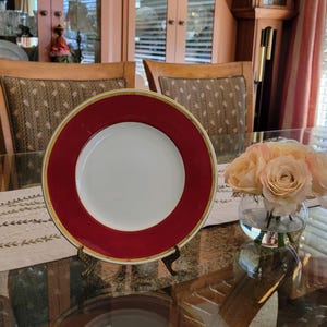 May include: A white plate with a red border and gold trim, displayed on a decorative stand. A small bouquet of peach-colored roses in a glass vase sits nearby. The plate is on a glass table.