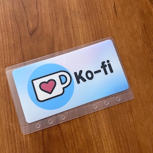 May include: A blue and white card with a coffee cup icon and the text "Ko-fi" printed on it. The card has a clear plastic cover and a row of holes along the bottom edge.