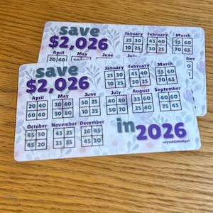 May include: Two cards with a floral design and the text "save $2,026 in 2026." Each card has a calendar layout with monthly savings goals. The cards are on a wooden surface.