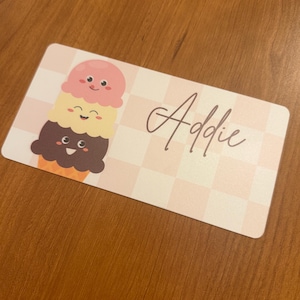 May include: A rectangular card with a pink and white checkered background. The card features a cartoon ice cream cone with three scoops in pink, yellow, and brown. The name "Addie" is written in cursive.