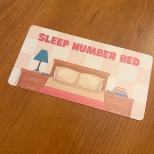 May include: A rectangular card with the text "SLEEP NUMBER BED" in pink letters. The card features an illustration of a bed with two nightstands, a lamp, and books. The background has a pink and white checkered pattern.