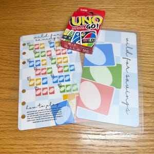 May include: A red and white UNO GO! card game box sits atop a clear plastic sleeve with colorful cards and a savings tracker. The tracker has the words "wild for savings" and "how to play" printed on it.