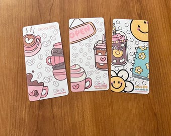 May include: Four colorful bookmarks with a coffee theme. Each bookmark features a different design, including a coffee cup, a coffee shop sign, a coffee drink, and a smiley face with a coffee cup. The bookmarks are all white with pink, blue, and yellow accents. The text "Save $7.5" is printed on each bookmark.
