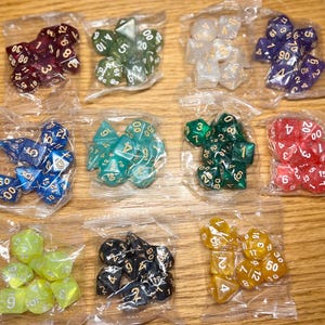 May include: A collection of polyhedral dice sets in clear plastic bags. The dice are in various colors, including red, green, blue, and yellow, with gold or white numbering. The sets are arranged on a wooden surface.