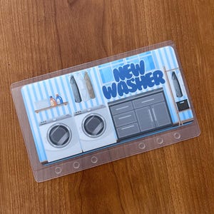 May include: A clear plastic card with a printed illustration of a laundry room scene. The image features two white washing machines, a gray cabinet, and the text "NEW WASHER" in blue and white.