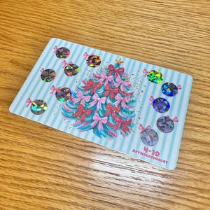 May include: A light blue card with a Christmas tree design. The tree is made of pink and blue bows and has a gold star on top. The card is decorated with holographic ornaments and the text "4-10 REYNOLDSBUDGET".