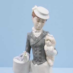 Op de afbeelding: Een porseleinen beeldje van een vrouw in een grijs jasje en witte rok die een klein wit hondje en een cadeaudoos met een blauw lint vasthoudt.