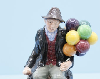 May include: A ceramic figurine of a man wearing a black coat and hat, sitting on a wooden bench and holding a bunch of colorful balloons.