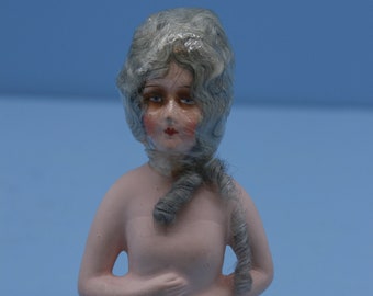 May include: A vintage porcelain doll head with a gray wig. The doll has painted features and is wearing a gray wig with a long, curled tail.
