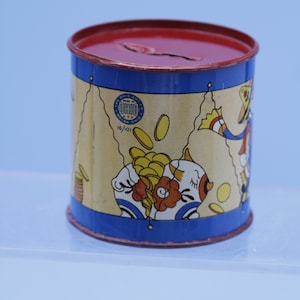 May include: A vintage blue and yellow metal piggy bank with a red lid. The bank features a cartoon pig with a red bow and a yellow hat. The bank has a circular sticker with the text "The Ohio Art Co. Bryan, Ohio 16/101".