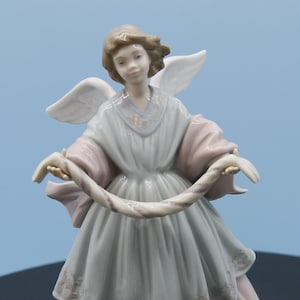 Könnte beinhalten: Porzellan-Engelsfigur mit einem ruhigen Ausdruck, trägt ein fließendes Kleid in den Farbtönen Hellblau und Rosa. Der Engel hält eine dekorative Girlande, mit weißen Flügeln und hellbraunem Haar. Die Figur ist etwa 25 cm groß.