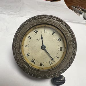 May include: Vintage round clock with a silver-coloured metal frame and a cream-coloured face. The clock has black Roman numerals and hands, with the text "WALTHAM U.S." visible. A textured knob is on the bottom.