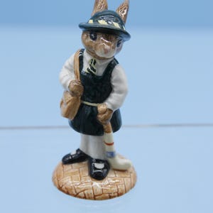 May include: A ceramic figurine of a rabbit wearing a green and yellow hat, a green and white school uniform, and holding a hockey stick. The rabbit is standing on a brown base.