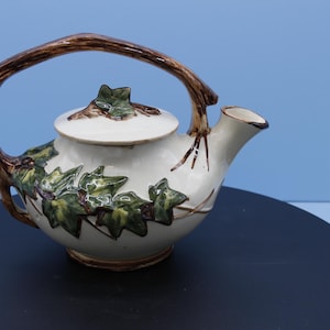 Könnte beinhalten: Eine Keramik-Teekanne mit cremefarbenem Korpus und braunem Griff. Die Teekanne ist mit grünen und braunen Efeublättern verziert. Der Deckel ist cremefarben mit einem braunen Blattmuster. Die Teekanne steht auf einer schwarzen Oberfläche.