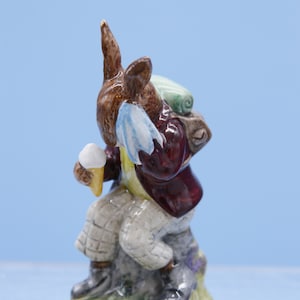 May include: A brown rabbit figurine wearing a red jacket, blue and white scarf, and green backpack, holding an ice cream cone. The rabbit is sitting on a rock with green grass.