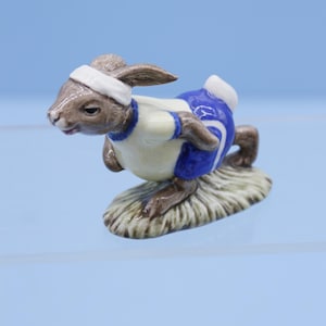 Puede incluir: Una figurita de cerámica marrón y blanca de un conejo que lleva un atuendo de correr azul y blanco. El conejo corre sobre una base verde.