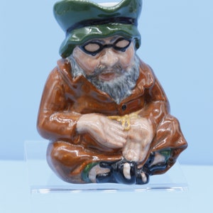 May include: A brown ceramic figurine of a man wearing a green hat and glasses. He is sitting with his hands clasped in his lap.