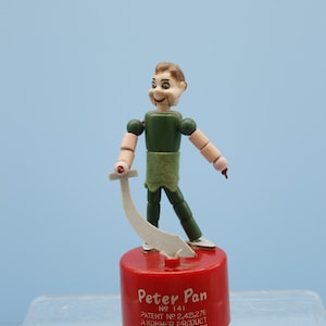 May include: A red and white Peter Pan finger puppet with a green shirt and a sword. The puppet is on a red base with the text "Peter Pan No 141 Patent No 2,421,279 A Kohner Product © Walt Disney Productions".