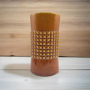 May include: A cylindrical ceramic vase with a glossy mustard-yellow finish. The upper and lower sections are smooth, while the central band features a raised, repeating grid pattern of small, pointed pyramid-shaped studs around the exterior.