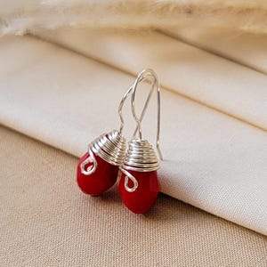May include: A pair of drop earrings featuring faceted red teardrop beads. Each bead is wrapped at the top with silver-colored wire that forms a small decorative loop, attached to a simple silver-colored hook for hanging from the earlobe.