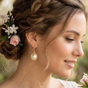 Puede incluir: Una persona con un peinado trenzado adornado con pequeñas flores blancas y rosas, que lleva un pendiente colgante dorado con una perla redonda y un top sin mangas de encaje blanco, sosteniendo un ramo de rosas blancas y rosas con flores pequeñas.