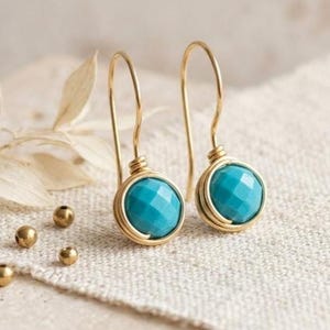 Puede incluir: Un par de pendientes de alambre dorado con cuentas redondas facetadas de color turquesa. Cada pendiente cuelga de un sencillo gancho dorado, mostrando una piedra azul brillante asegurada por un delicado marco de alambre dorado.