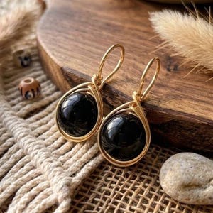 Puede incluir: Un par de pendientes colgantes con piedras negras redondas y pulidas, engastadas en marcos de alambre de tono dorado. Los pendientes tienen cierres de gancho simples en la parte superior, diseñados para colgar verticalmente con brillo.