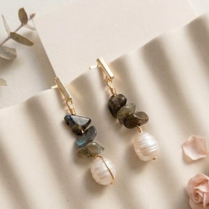 Puede incluir: Un par de pendientes colgantes dorados con un poste de barra vertical, una serie de piedras de labradorita irregulares de color gris verdoso oscuro y una perla de agua dulce blanca de forma ovalada en la base, unidas por un hilo dorado.