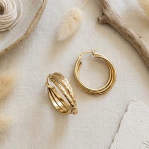 May include: A pair of gold-toned hoop earrings, each featuring a triple-layered design. One hoop displays a smooth, polished finish, while the other two hoops exhibit a textured, hammered surface. The earrings are circular with a hinged clasp.