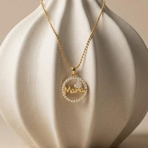 Puede incluir: Un collar de tono dorado con un colgante circular con un borde de cristales engastados. El centro del colgante muestra la palabra Mama en letra cursiva, acompañada de un pequeño corazón incrustado de cristales sobre el texto.