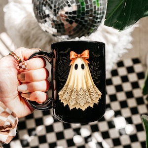 May include: Black ceramic mug featuring a whimsical ghost design. The ghost is draped in a cream-colored sheet with a lace trim and an orange bow. The mug has a black handle and is decorated with bats and a spiderweb.