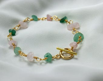 May include: A gold-toned chain bracelet featuring alternating raw, tumbled light pink and green stones. The bracelet is secured with a gold-toned toggle clasp consisting of a circular ring and a bar, with each stone individually wire-wrapped.
